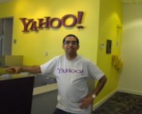 Young man, dark skin, short dark hair, wearing white t-shirt with word YAHOO!, hand on hip, standing in office with yellow walls and YAHOO! sign.