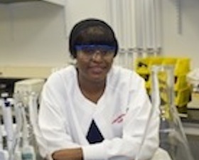 Young black woman, wearing white lab coat, leaning on lab table in lab room environment.