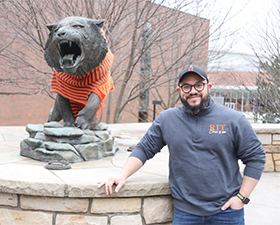 Photo of Arturo Peralta Silva, young man wearing RIT shirt and baseball cap, hand in pocket, posing by the RIT tiger statue wearing an orange stripped sweater.