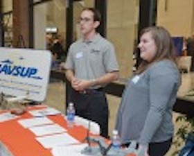 Young man, short dark hair, glasses, grey shirt, standing at corporate presentation table with a colleague, sign says NAVSUP.