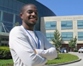 Young man, dark skin, wearing white top, arms crossed over chest, standing in front of building with words Rebecca and John Moores UCSD Cancer Center.