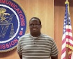 Young man, dark skin, glasses, striped shirt, hands crossed behind back, standing in front of  Federal Communications Commission plaque and American flag.