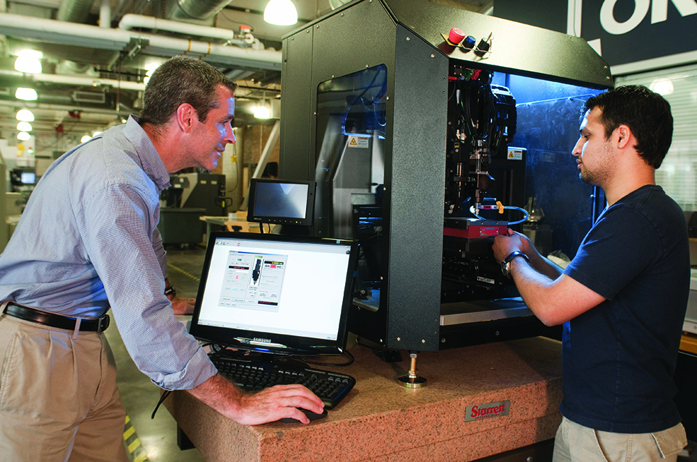 Denis standing over a laptop while peering into som 3d printer with another person working beside him