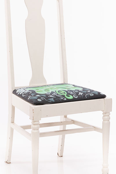 White chair with a painted cushion. The painting on the cushion is of a lunar moth on a dark navy background with various organic and plant like shapes. 