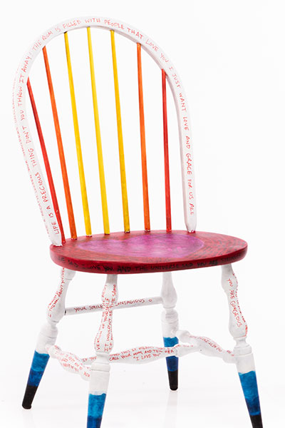 The legs have a black-blue-white gradient, the seat is red with a pink heart in the middle with the words "feel the love" written on it, the pillars to the backing are a gradient from yellow to red-orange from the middle out, the backing arch is white. There are phrases written all over the chair in red, blue and black.
