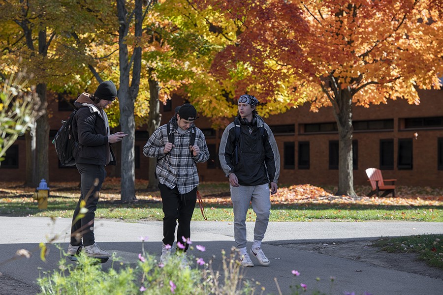 3 male students walking together on an outdoor path in the Fall. Foliage visible in the back. 