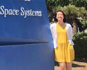 Photo of Amanda Bui, young woman with dark hair, wearing yellow dress and white sweater, standing by Lockheed Martin sign.