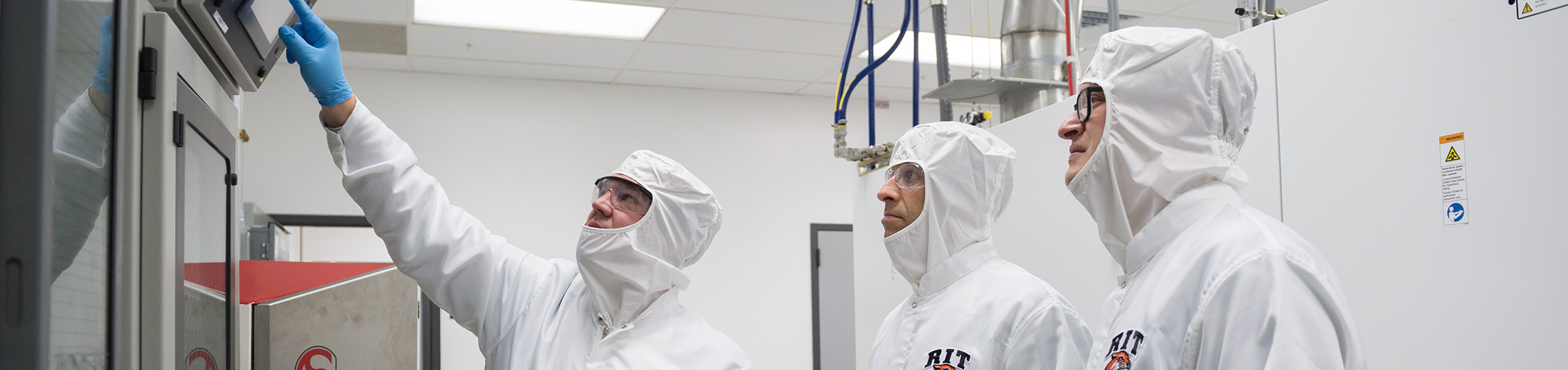 3 people in a clean room wearing white protective suits.