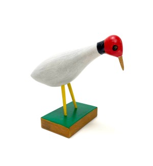 a carved wood bird with a red head and white body, black stick legs on a green block stand.