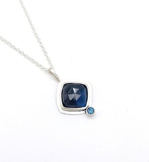 a Sterling silver chain with a pendant with a dark blue faceted stone set in a flat silver bezel setting with a small light blue topaz at the tip.