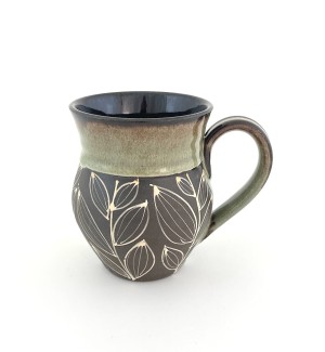 a hand thrown ceramic mug with a dark clay body, illustrated with a stylized leaf pattern and a green glaze at the rim.