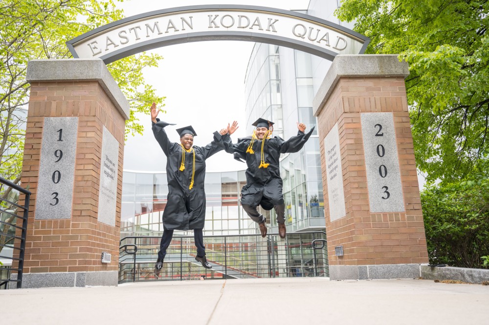 two students jump joyfully in front of a sign for the Eastman Quad.