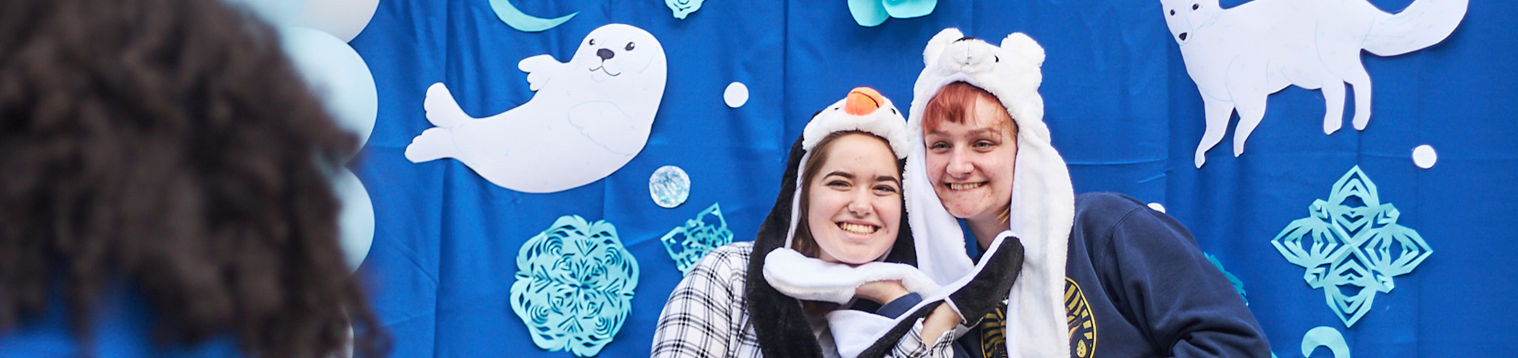 Two students pose for a picture against a blue FreezeFest-themed backdrop featuring cut outs of seals, foxes, and snow flakes