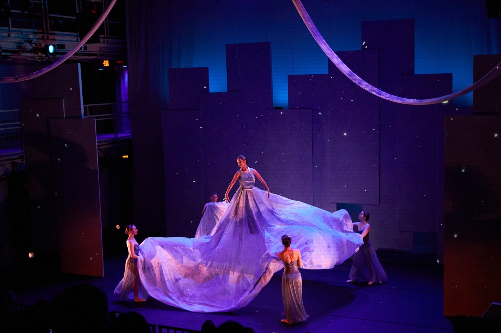 a dancer on a stage, raised several feet in the air, with a long and wide skirt ballooned out and held by four other dancers.