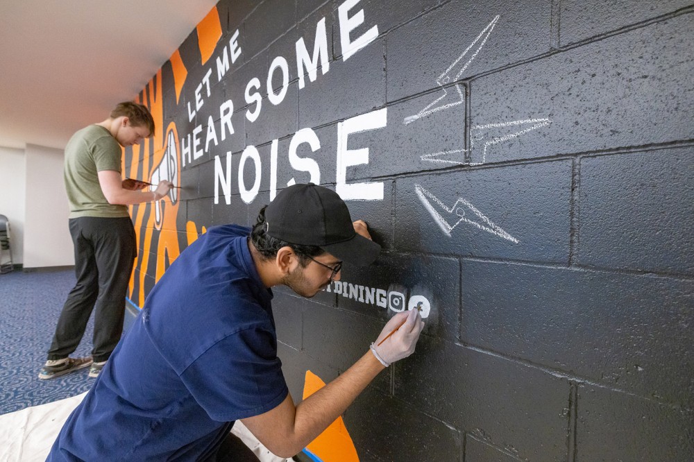 wo artists paint a vibrant mural on a black brick wall, featuring bold white text reading 'Let me hear some noise' alongside graphic elements and social media icons.