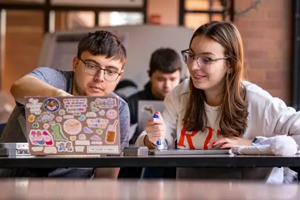 A tutor helping a fellow student, while sitting at a table in front of a laptop.