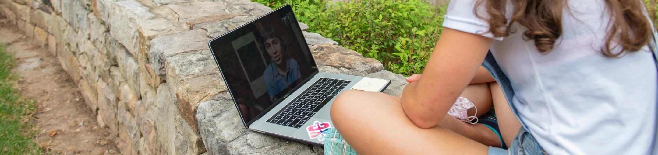 An RIT student sitting on a stone wall talking to someone via video conference software.