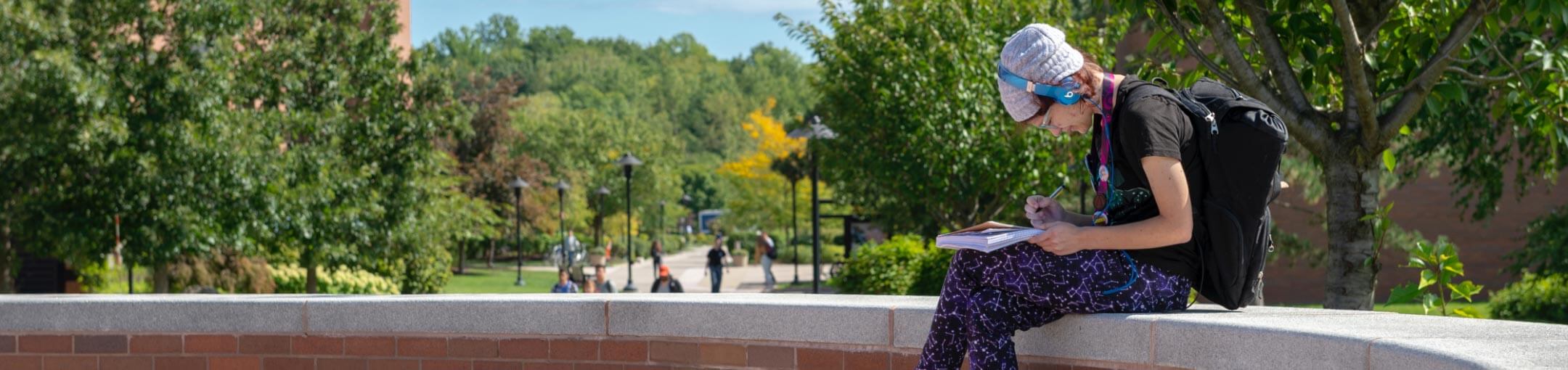 An RIT student sitting on a wall in a quad, writing in their notebook.