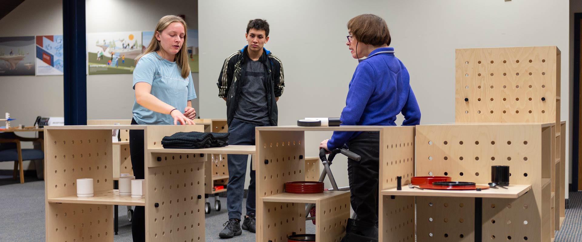 two students describe a modular kitchen cabinet system to a person using a walker.