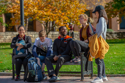 A group of diverse students standing and sitting on a bench talking on RIT campus.