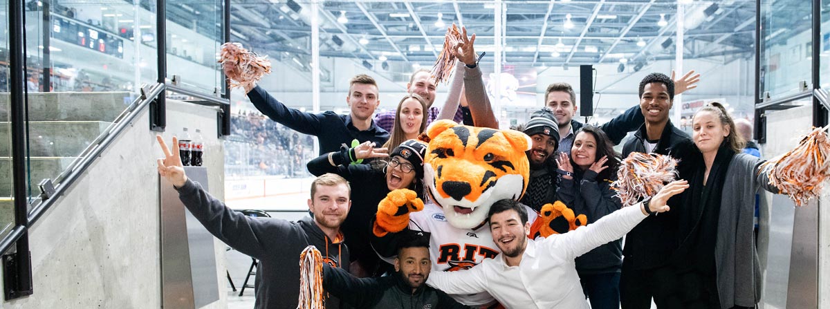 students stand with the RIT mascot in front of an ice rink