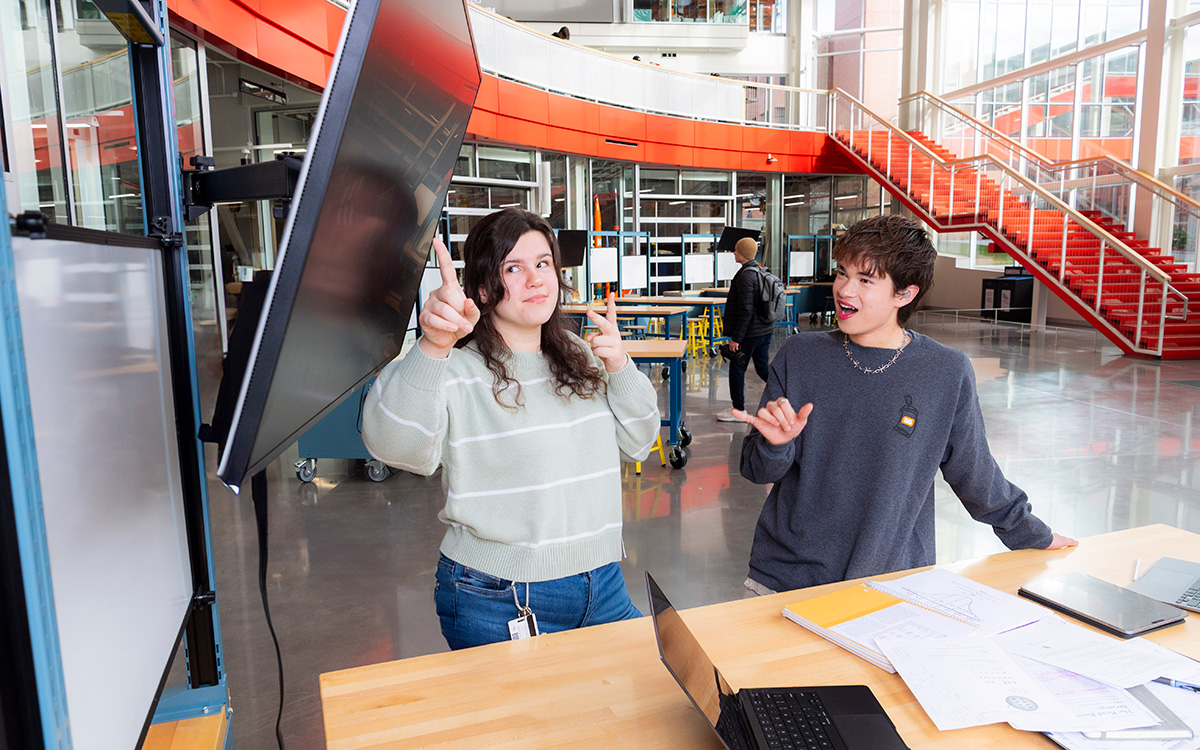 Two students signing A S L and pointing at a screen