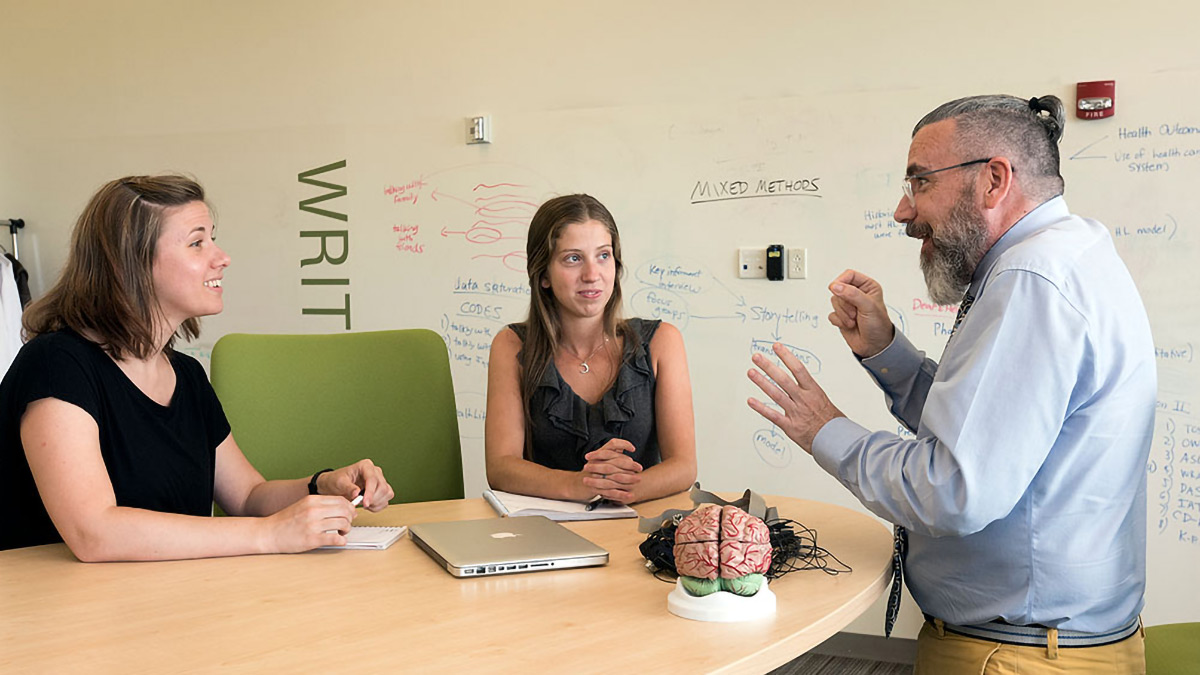 Three people in discussion, one using sign language, at a round table with notes and a model brain on display.