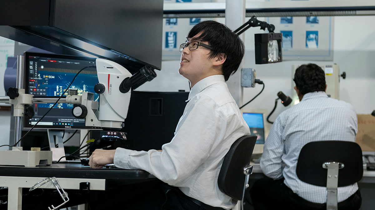 Two individuals working in a lab, one observing circuit boards through a microscope.