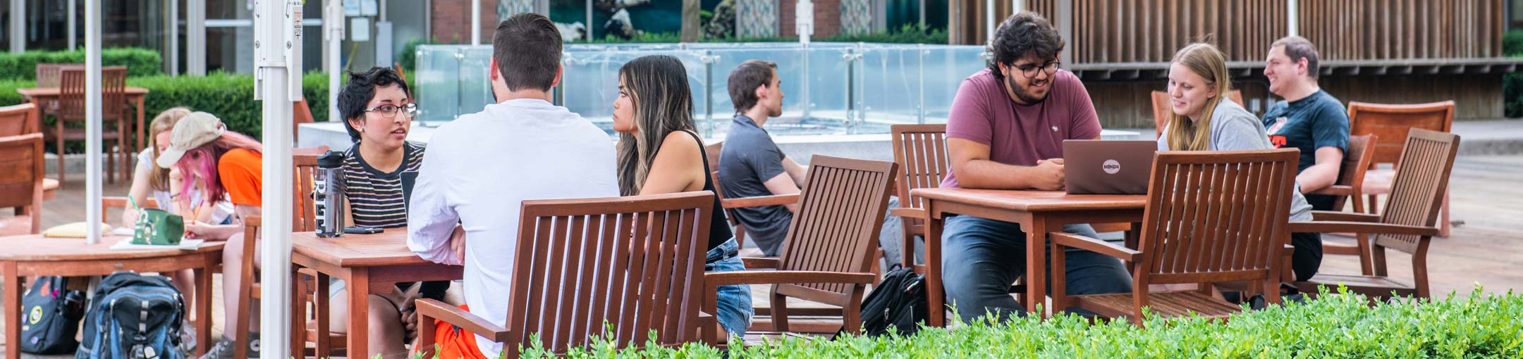 students sit at tables in a plaza by a water feature