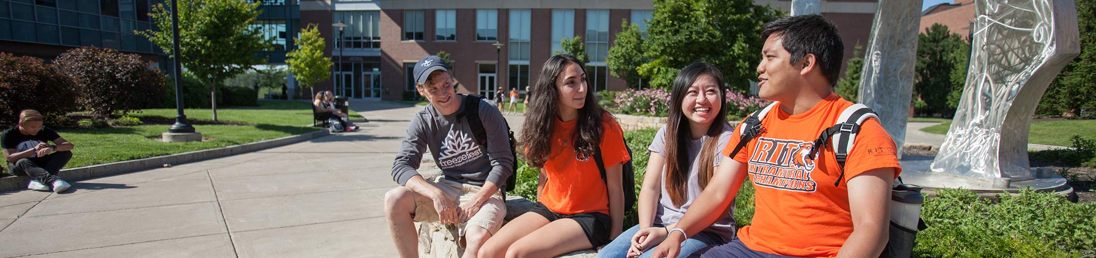 Students sitting on a brick wall in a courtyard on a sunny day wearing orange RIT shirts