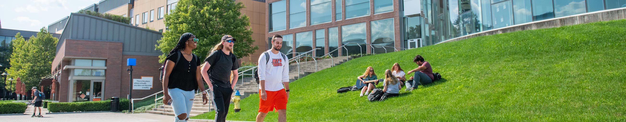 a group of students walks past another group of students who are sitting on a lawn on the RIT campus