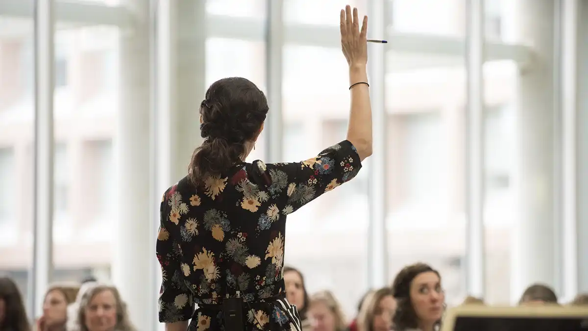 the back view of a woman addressing a group in a meeting space, with her arm raised.