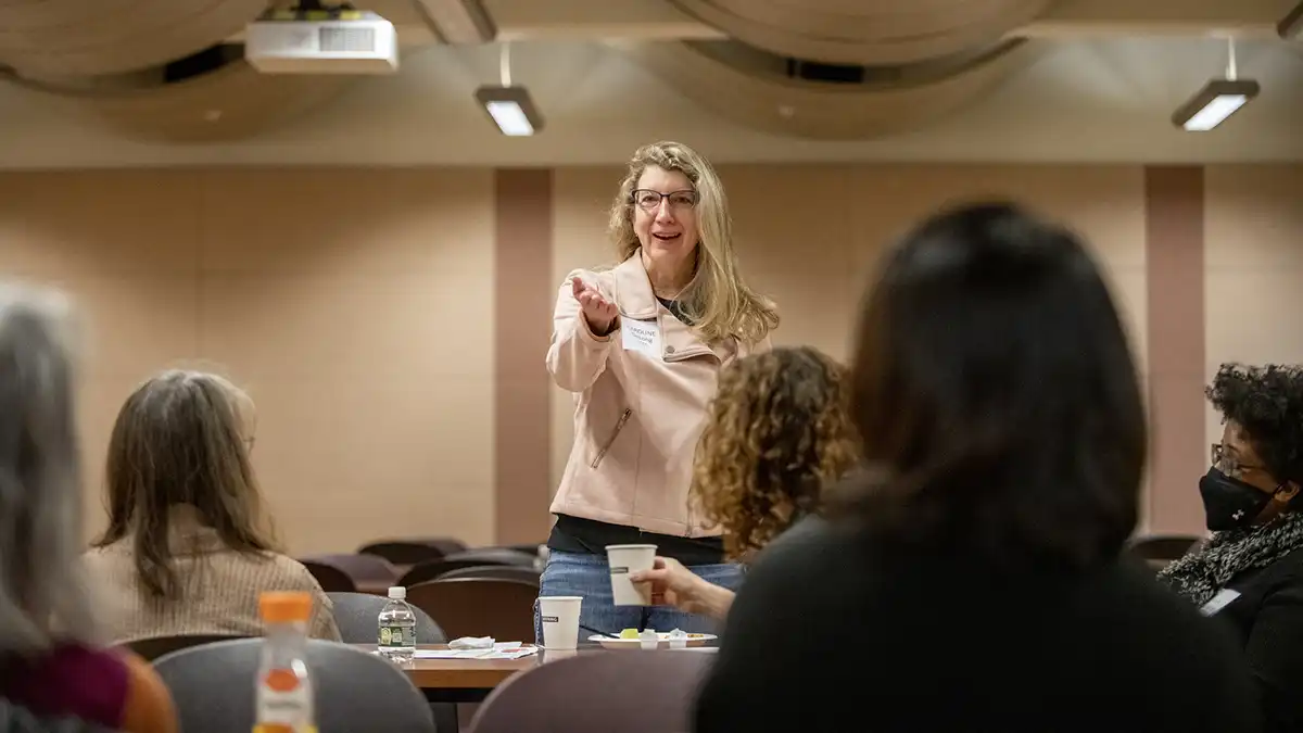 a woman gesturing as she gives a presentation in front of tables of participants.