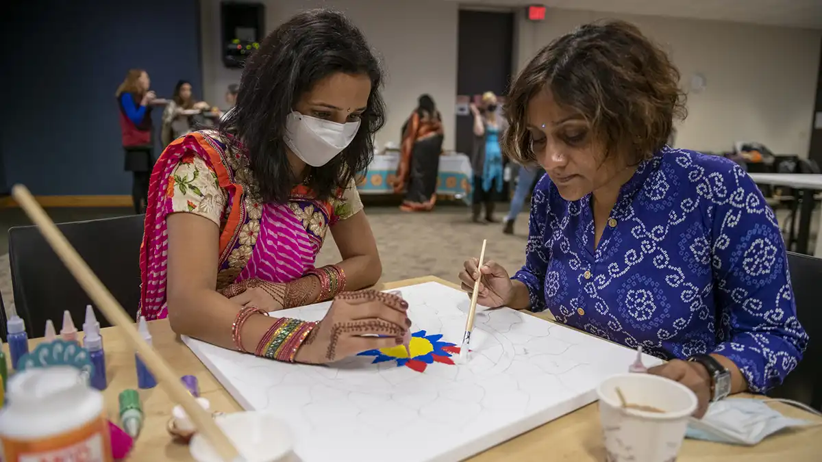 two women dressed in traditional Indian clothes working on a colorful design piece.