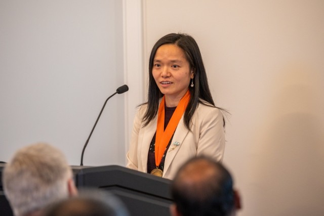 Woman wearing orange medallion stand at podium in front of microphone.