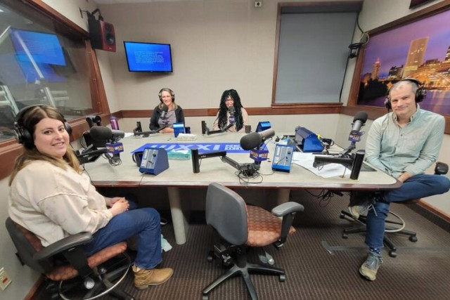 Four people wearing headphones sit at a table in a radio talk studio: a woman front left has long brown hair and is wearing a beige shirt, blue jeans and brown boots; a man front right has short dark hair and is wearing a light green button-down shirt, blue jeans and sneakers; a woman back left has long blonde hair and is wearing a black zip-up fleece over a grey t-shirt; a woman back right has long black and green dreadlocks and is wearing glasses and a pink shirt.
