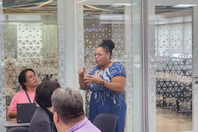 a woman in a blue and white dress speaks to other adults in a modern classroom environment.