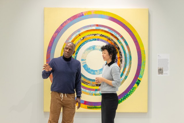 a man and a woman stand in an art gallery, with the man gesturing as explaining a piece of art beyond the field of view.