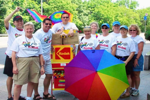 A group of men and women hold rainbow umbrellas and wear T-shirts reading 'Pride @ Kodak.