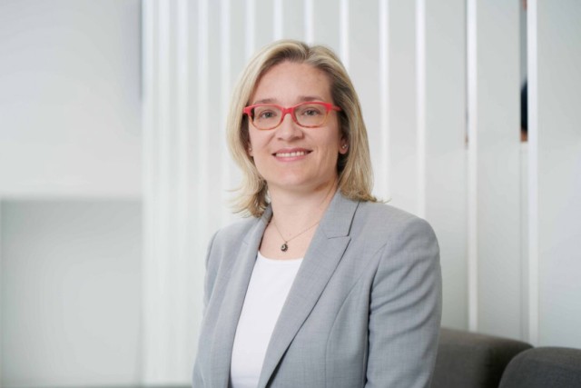 a woman with blonde hair and red glasses wears a gray suit and poses for a photo in a white room.
