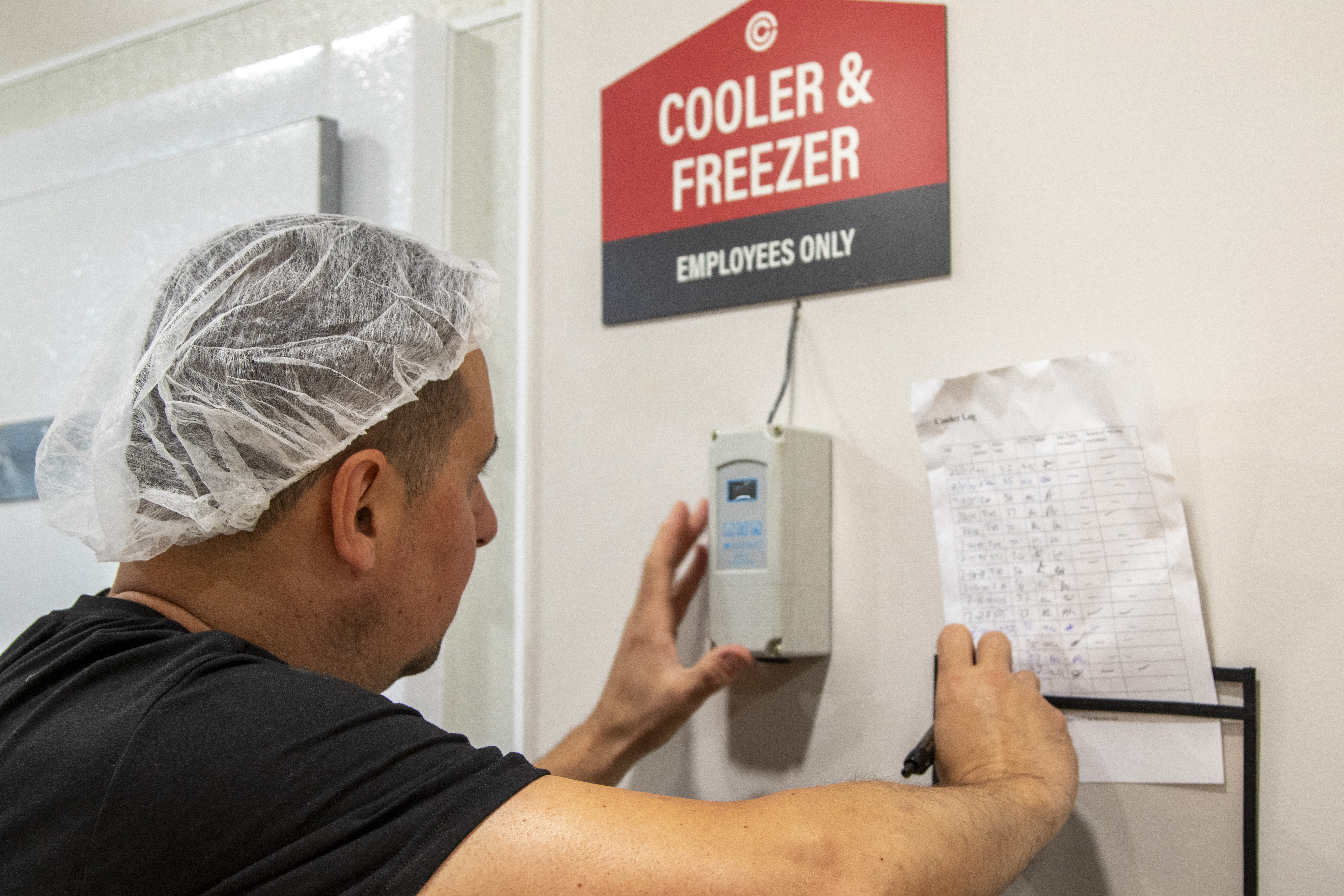 worker wearing hairnet writing down temperature readings on a piece of paper hanging outside of a cooler/freezer