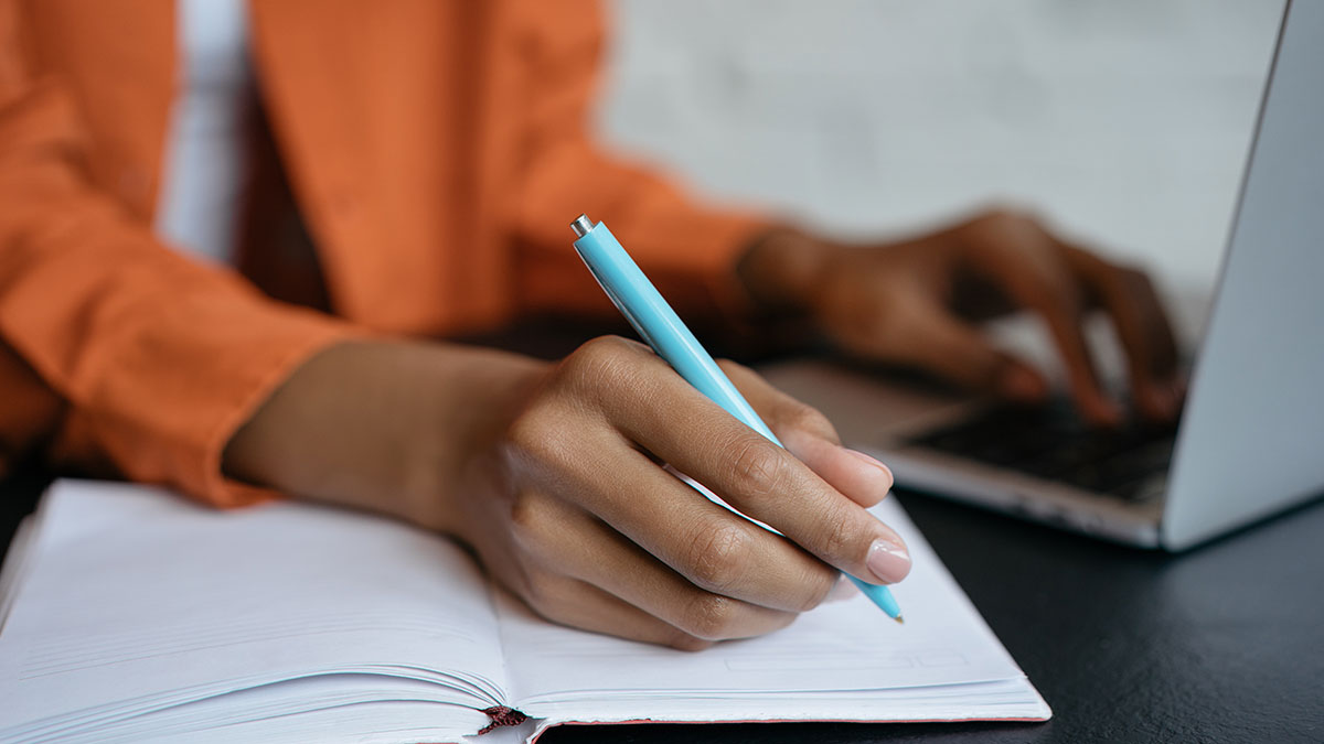 Close-up of a person writing in a notebook with one hand while using a laptop with the other.