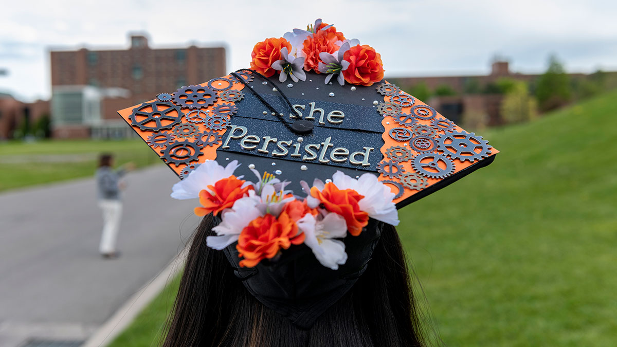 A decorated graduation cap with orange flowers and gears reads 'she Persisted' in gold letters.