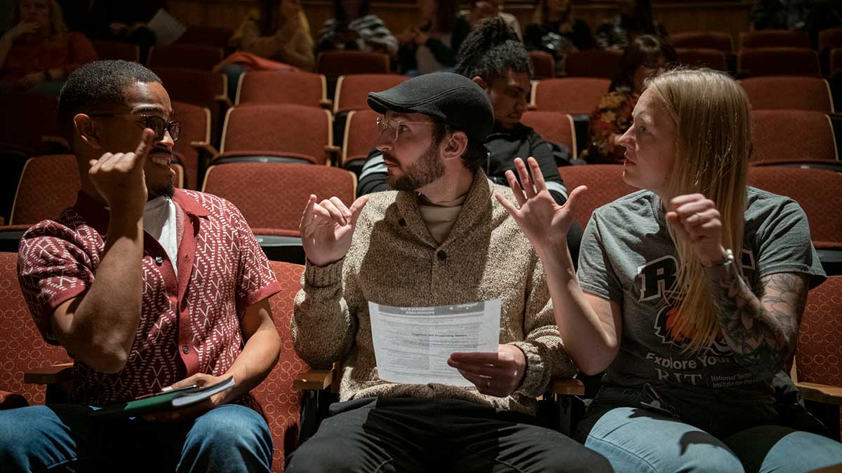 Three people sit in a theater's audience seats conversing using sign language.