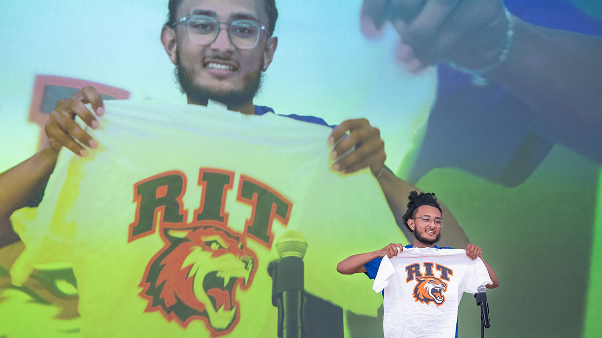 A student holds up an R I T Tigers T-shirt on stage in front of a large projected image of himself.