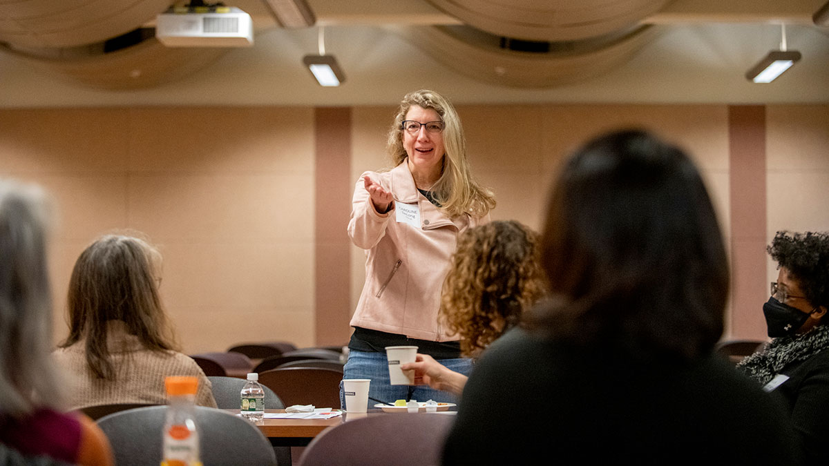 A woman speaks and gestures to a seated group during an indoor workshop.