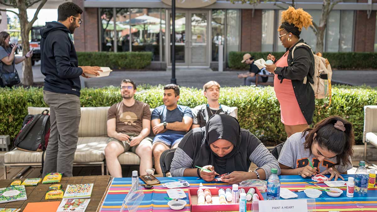Students create art at a colorful outdoor table while others chat nearby.