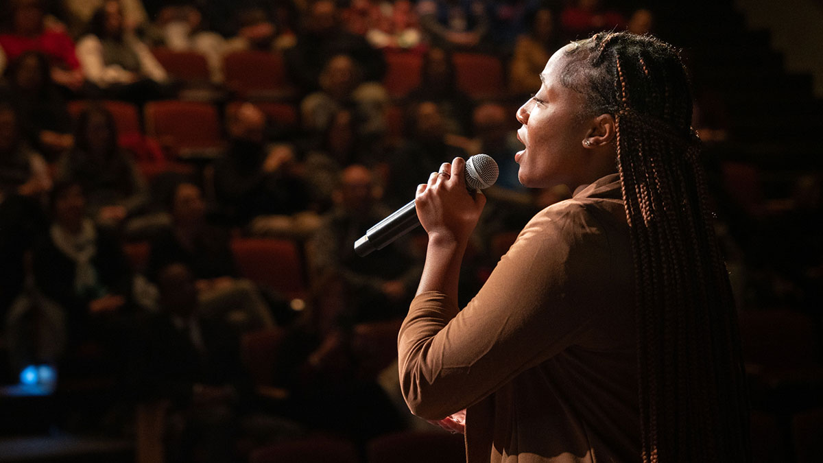 A young woman sings into a microphone on stage in front of a blurred audience.