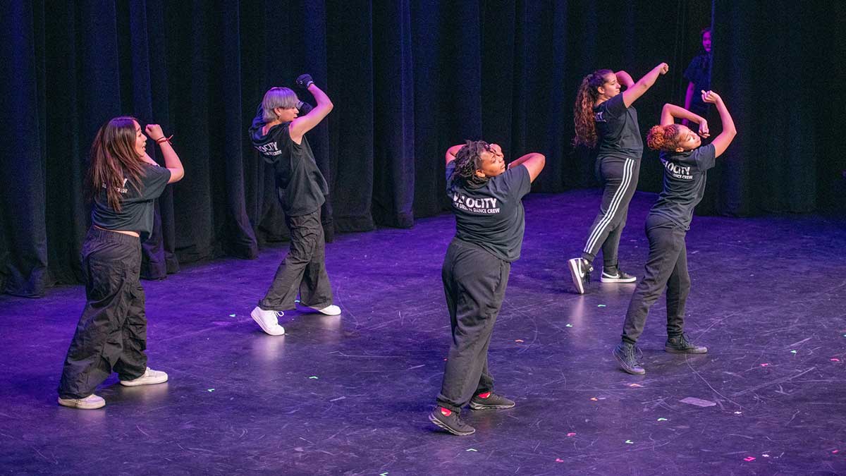 Five dancers perform synchronized moves on a stage wearing matching black shirts.