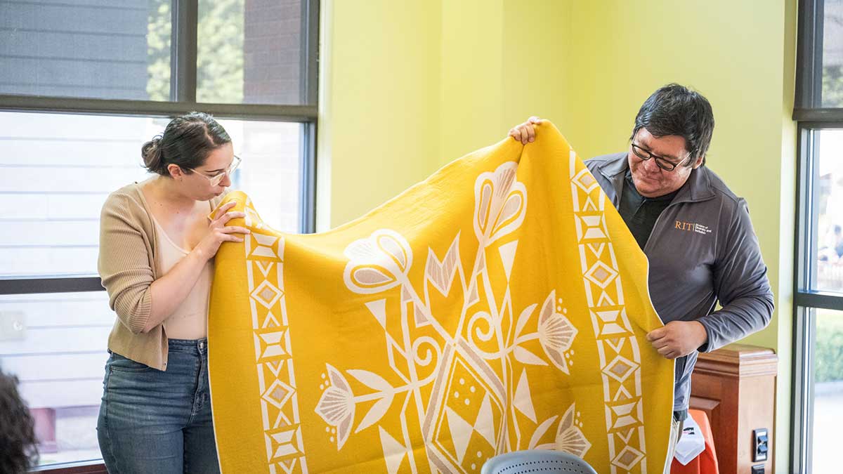 Two people hold up a large yellow blanket with white floral and geometric designs in a classroom.